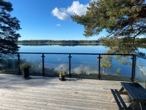 Holzterrasse mit schwarzem Pfosten-Glasgeländer, das ein Seegrundstück umrahmt und freien Blick auf Wasser und Natur bietet.