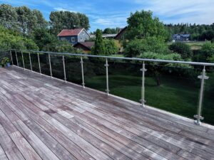 Balkon­geländer mit getöntem VSG und Edelstahlpfosten auf Holzterrasse mit Blick auf Garten und modernes Einfamilienhaus.