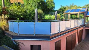 Terrasse mit mattiertem Glasgeländer und runden Edelstahlpfosten, umgeben von grünem Garten und Wintergarten.
