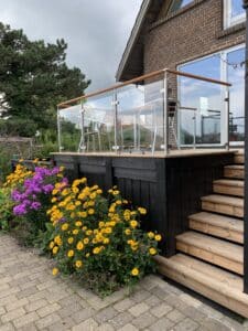 Edelstahl-Glasgeländer mit Holzhandlauf auf einer Terrasse mit schwarzer Holzverkleidung und bunten Blumen im Vordergrund.