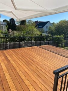 Schmiedeeisernes Geländer in Schwarz auf Holzterrasse mit Markise, sonnenbeschienenen Dielen und grüner Gartenumgebung.