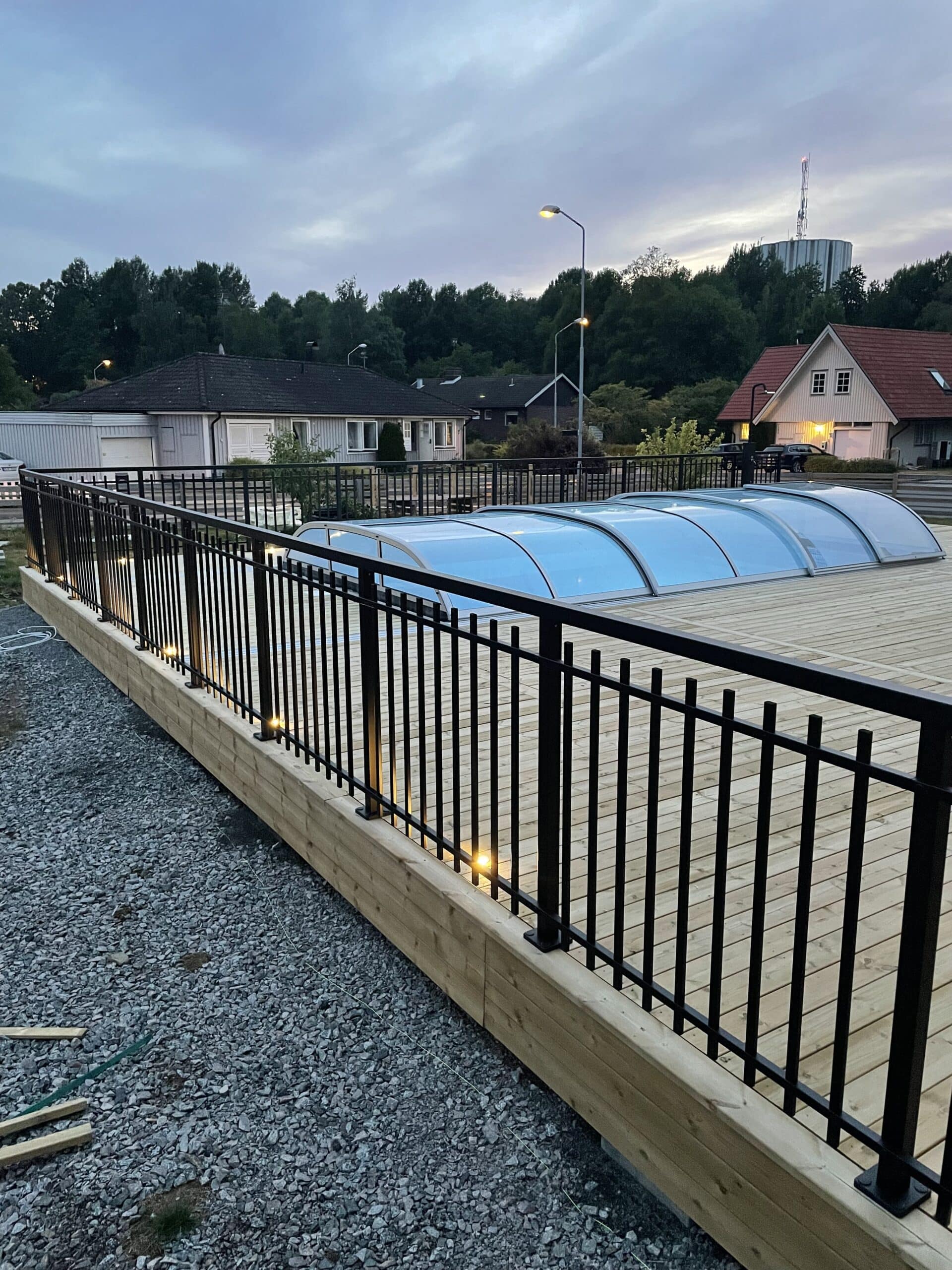 Schwarzes Schmiedegeländer aus Aluminium von Geländerladen rund um einen Pool mit Holzterrasse, blauer Poolabdeckung und integrierter Beleuchtung bei Abendlicht