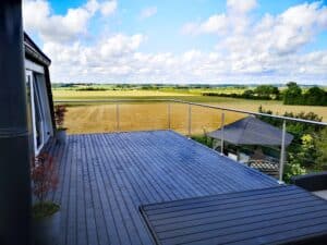 Edelstahlgeländer mit Drahtseilsystem auf Terrasse mit dunkelgrauen Dielen, mit freiem Blick auf Äcker und grüne Felder.