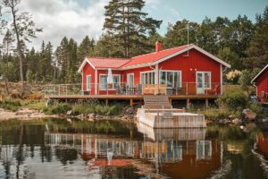 Rotes Seehaus mit umlaufendem Glasgeländer, Terrasse mit Treppe zum Steg und ruhiger Wasserfläche im Vordergrund.