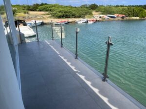 Glasgeländer mit Edelstahlpfosten, montiert entlang eines Stegs mit Blick auf einen Yachthafen und grüne Landschaft im Hintergrund.