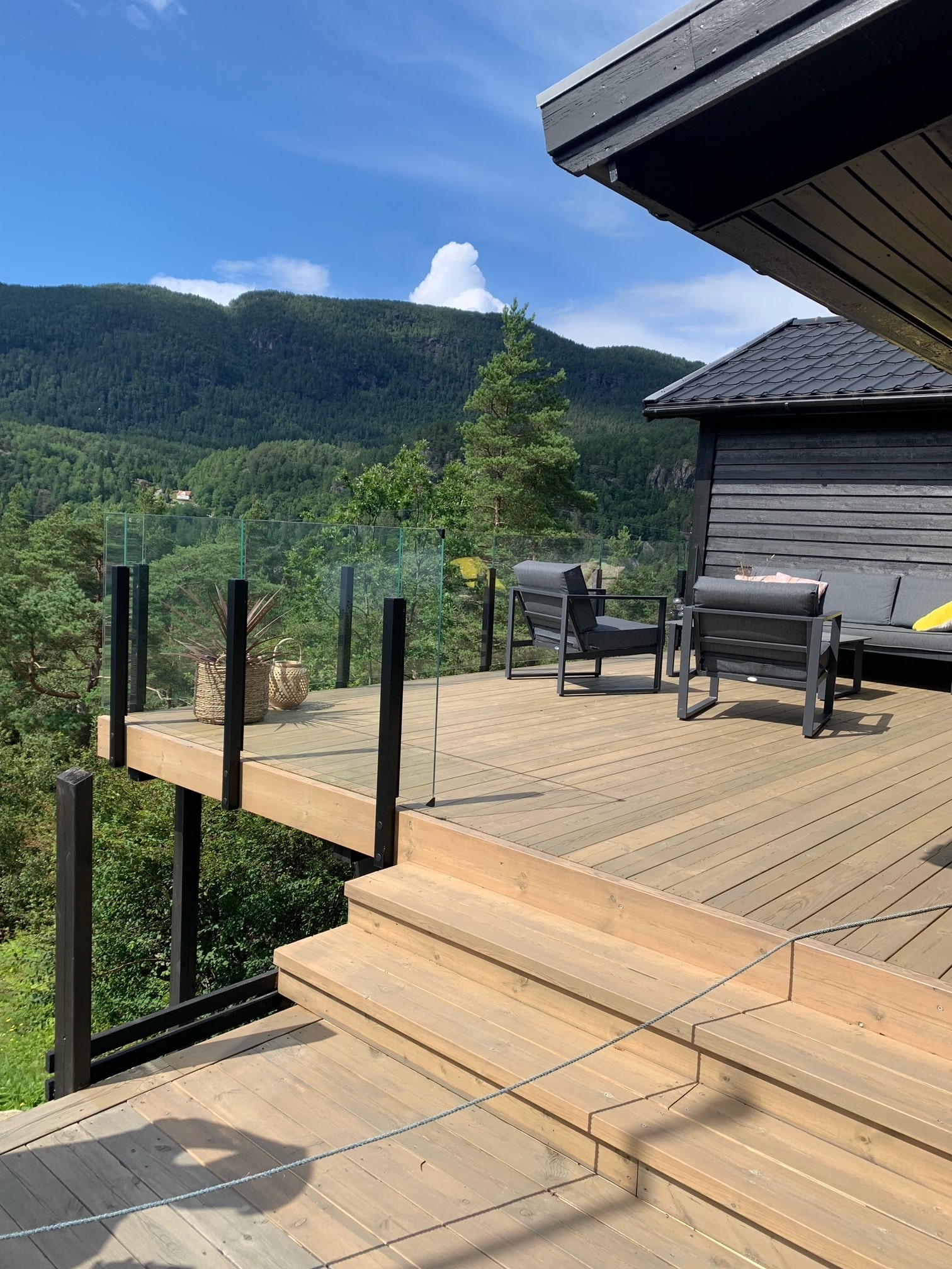 Terrasse mit schwarzem, pfostenlosem Glasgeländer mit Ausblick auf Wald, Berge und blauen Himmel
