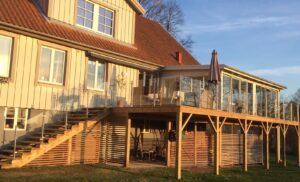 Glasgeländer aus Edelstahl und Klarglas, montiert entlang einer Treppe und mehrstufiger Holzterrasse mit Wintergarten und sonniger Fassade.