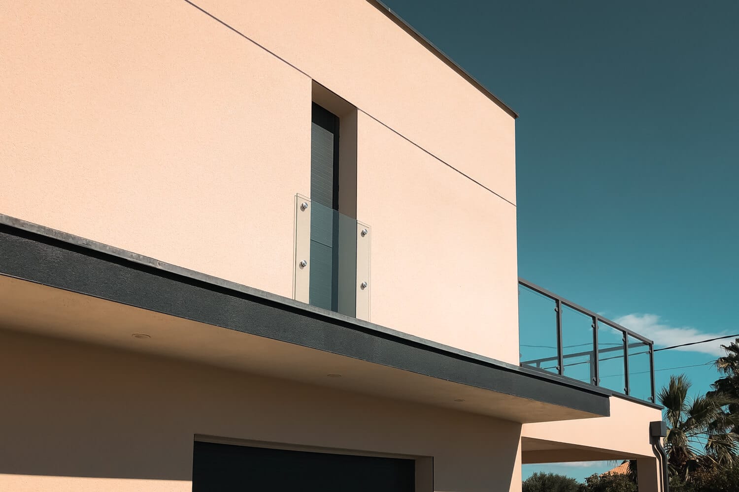 Französischer Balkon aus Klarglas mit Fixpunkten vor der Terrassentür eines modernen Hauses mit verputzter Fassade und schwarzer Dachkante