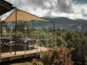 Glasgeländer auf mobiler Terrasse mit Bergpanorama Höhenverstellbare Terrasse mit Glasgeländer, Edelstahlpfosten und Sonnensegel – auf Rädern montiert mit Blick auf grüne Täler & Alpen.