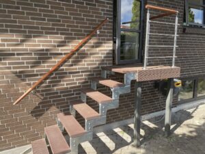 Verzinkte Stahltreppe mit Holzhandlauf und Edelstahl-Drahtseilen. Der Wandhandlauf folgt einer roten Backsteinfassade und verbindet Stabilität mit modernem Stil.