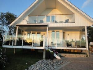 Glasgeländer für Balkon und Terrasse Weißes Holzhaus mit Glasgeländern auf Balkon und Terrasse – Edelstahlpfosten, Klemmbefestigung und warme Abendsonne im Glas reflektiert.
