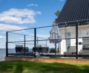 Schwarzes Aluminiumgeländer mit Glas Terrasse mit schwarzem Aluminium-Glasgeländer auf Holzdeck neben weißem Haus – Seeblick, blauer Himmel und moderner Windschutz.