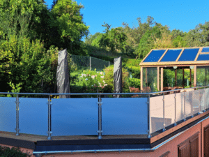Edelstahlgeländer mit satiniertem Glas Balkon mit Edelstahlpfosten, Milchglas und runder Handlauf – montiert auf erhöhter Terrasse mit Garten, Glashaus und blauer Himmel.