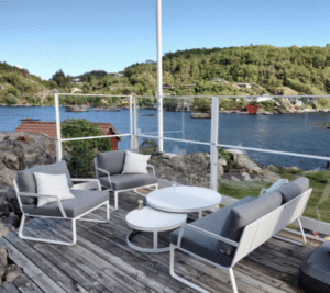 Aluminium-Glasgeländer mit Meerblick Terrasse mit weißem Aluminiumgeländer auf Felsvorsprung am Meer – glasklare Sicht auf Bucht, rote Bootshäuser und grüne Hänge.