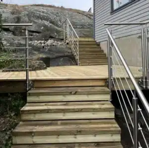 Holztreppe und Balkon mit druckimprägniertem Belag und Edelstahlgeländer mit horizontalen Stäben. Die moderne Fassade und Bergkulisse runden das Bild ab.